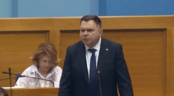 Političar u tamnom odijelu sa plavom kravatom govori za govornicom u skupštinskoj sali.