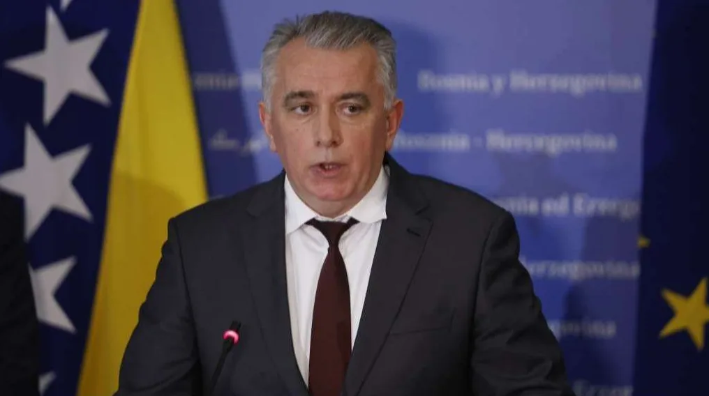 Političar u odijelu s tamno crvenom kravatom, pored zastave Bosne i Hercegovine.