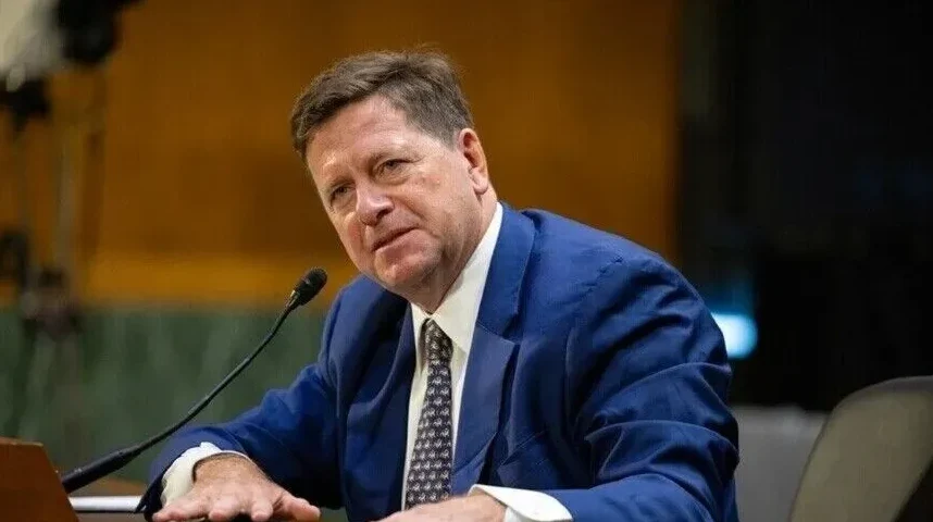 Portret muškarca u plavom odijelu i bijeloj košulji sa kravatom, koji sjedi za stolom i govori za mikrofonom.