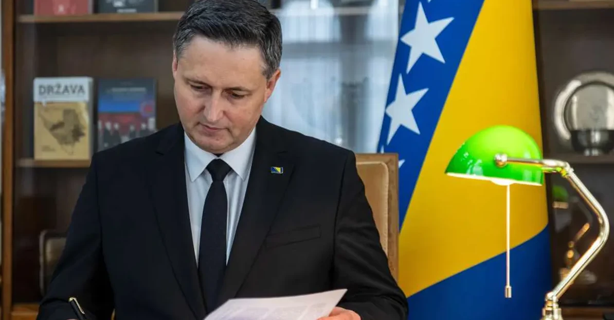 Političar u crnom odijelu, sa značkom zastave Bosne i Hercegovine na reveru, sjedi za stolom i potpisuje dokument, pored njega je zastava Bosne i Hercegovine i zelena stona lampa.