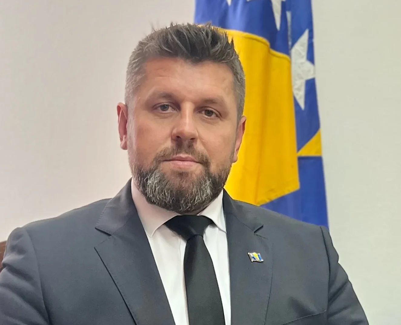 Portret potresenog muškarca u tamnom odijelu i bijeloj košulji s crnom kravatom, s Kosovskom zastavom u pozadini.