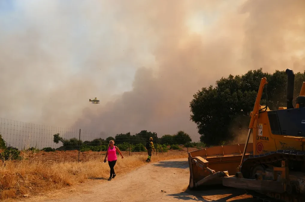 Avion za gašenje požara leti iznad gustog dima, dok se vatrogasac i buldožer nalaze na zemlji.
