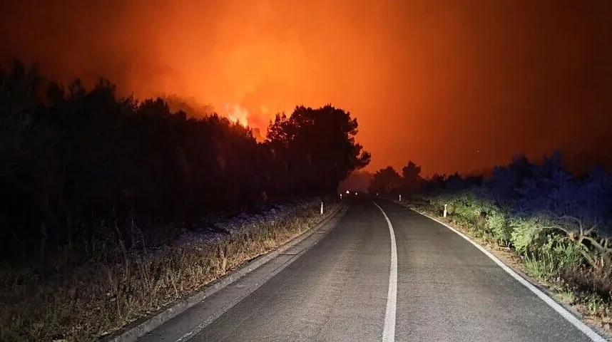 Put noću osvijetljen farovima automobila, sa gustim dimom i narandžastom vatrom u pozadini.