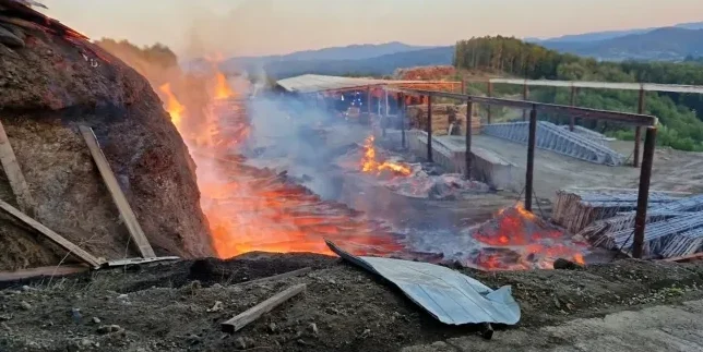 Veliki požar bukti u pilani, sa plamenovima koji izbijaju iz hrpe drveta i dima koji se diže u zrak.