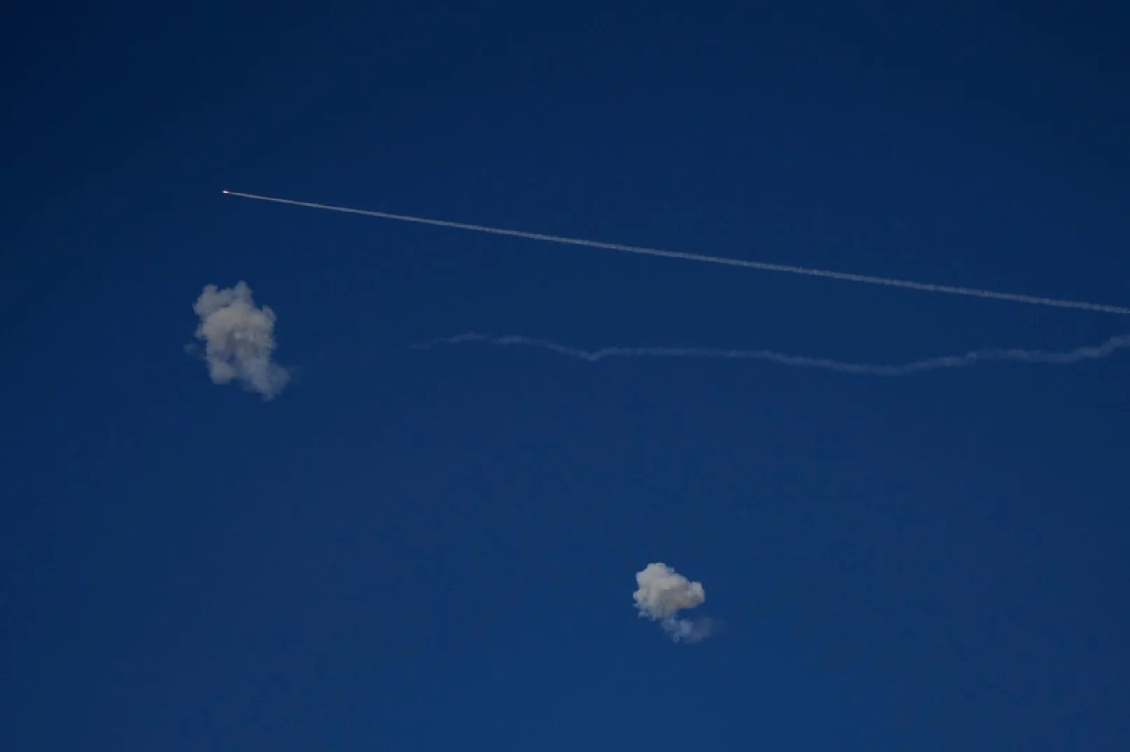 Plavi nebeski svod sa dugim, bijelim tragom aviona ili rakete koji se proteže od gornjeg lijevog do desnog ugla, uz jednu oblak dima u sredini lijevo i drugi manji oblak dima u donjem desnom uglu.