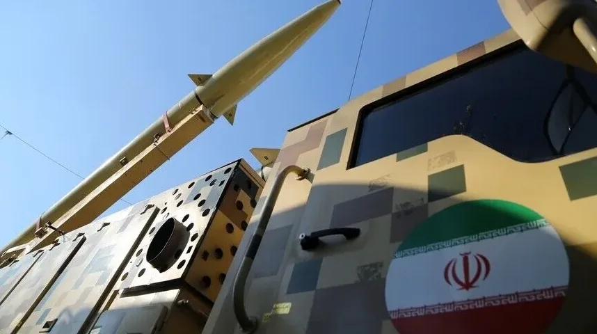 Detaljan pogled na lansirnu cijev rakete, koja je usmjerena prema nebu, iznad kamuflažno obojenog vozila. Na vozilu je jasno vidljiva iranska zastava s natpisima na arapskom jeziku i simbolom Allaha.