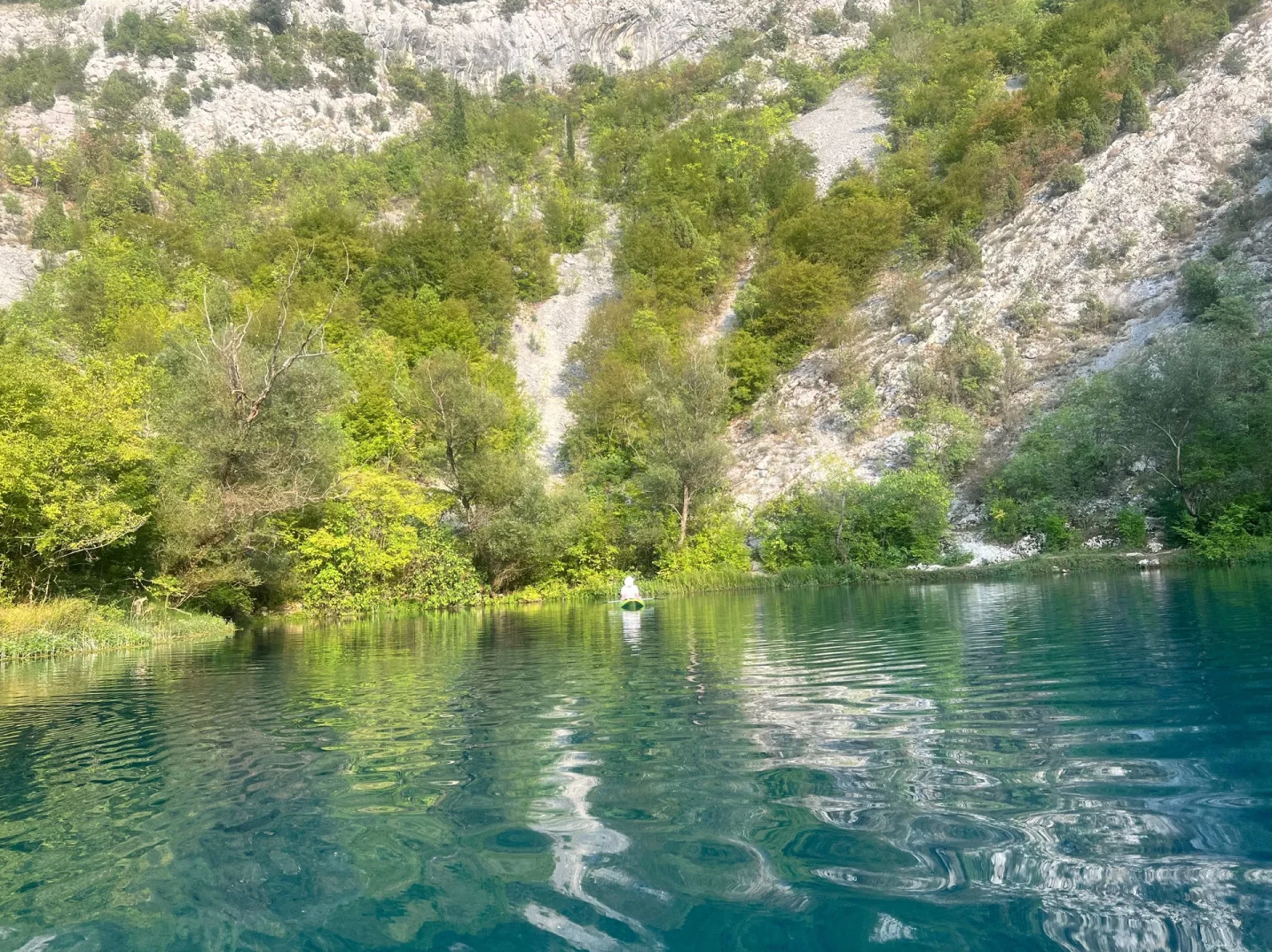 Čovjek vesla na kajaku po bistroj tirkiznoj rijeci, okruženoj bujnim zelenilom i kamenitim padinama.