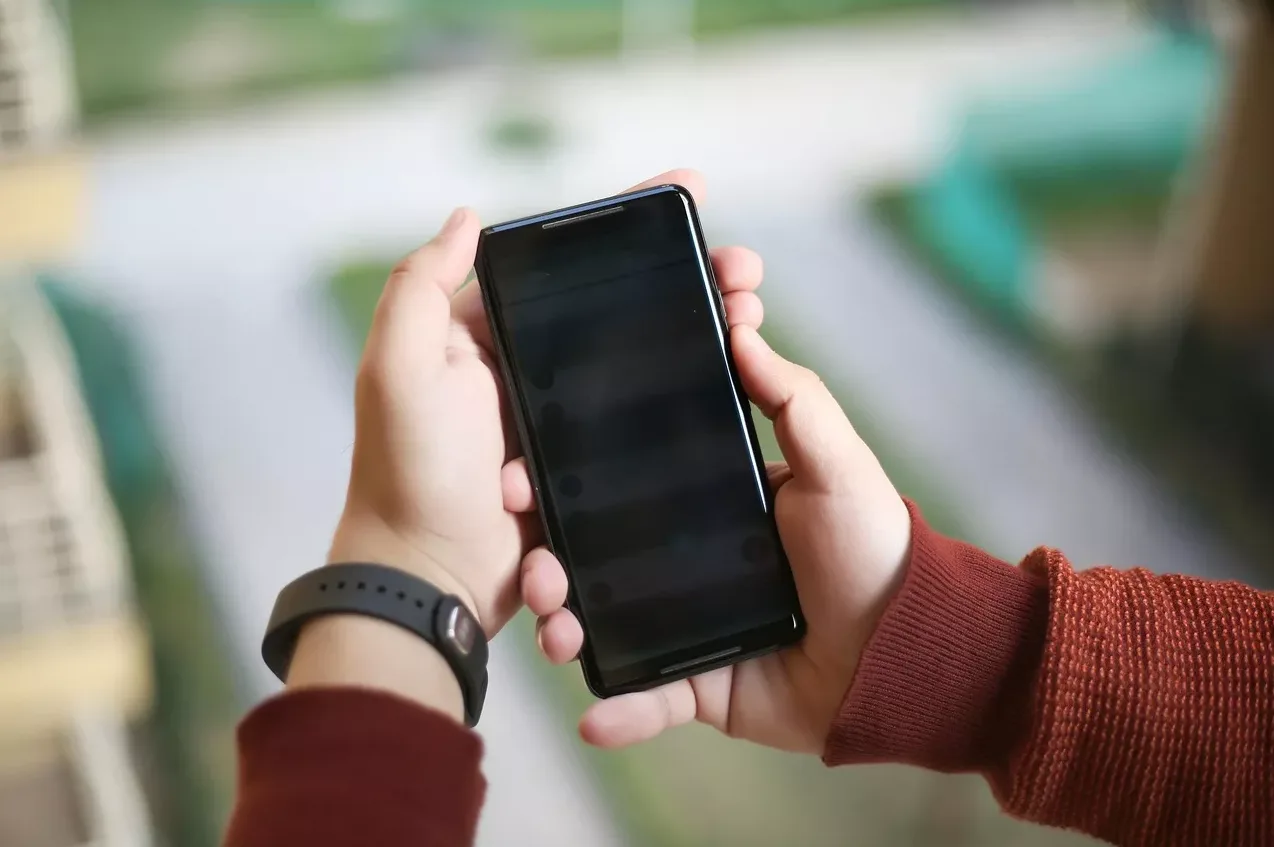 Osoba drži crni mobilni telefon u rukama, a na lijevoj ruci nosi crnu pametnu narukvicu.