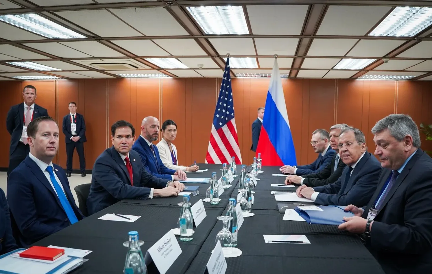 Američka i ruska delegacija sjede za stolom na sastanku, sa zastavama SAD-a i Rusije u pozadini.