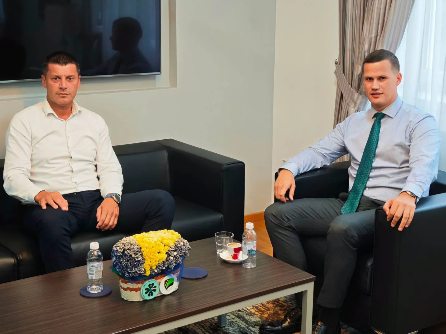 Dva poslovna čovjeka sjede na crnim kožnim garniturama u moderno uređenoj kancelariji.