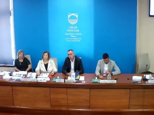 Četiri osobe sjede za stolom ispred plavog panoa s logom Grada Mostara.