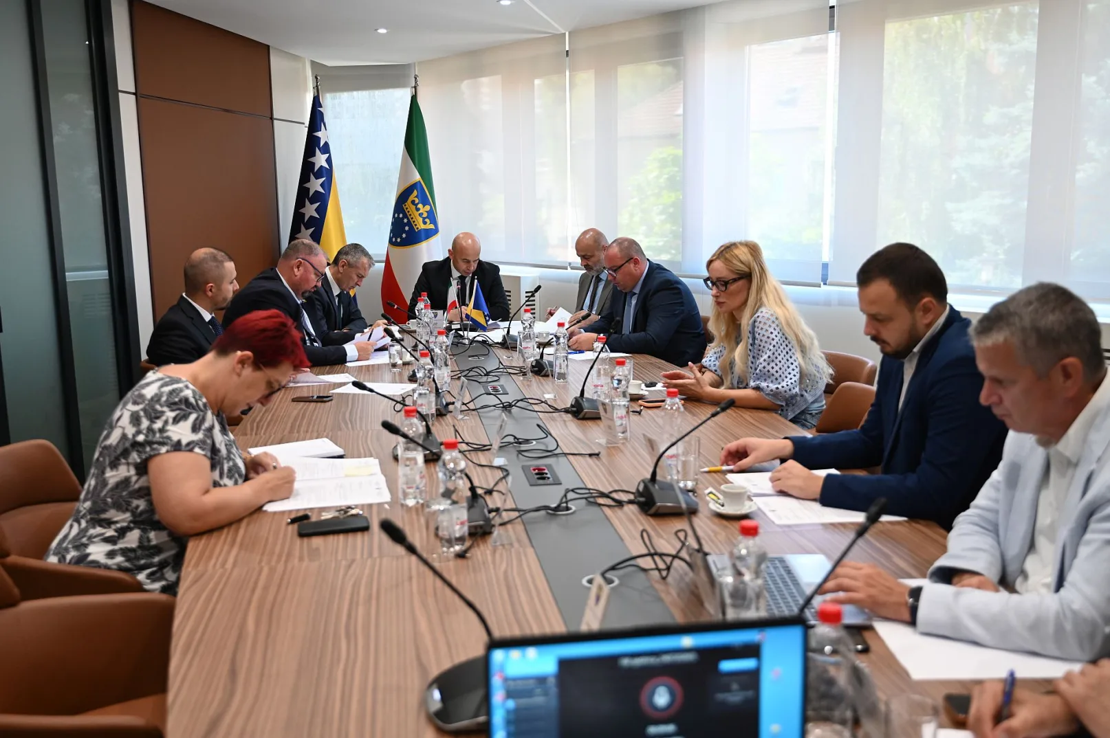 Grupa ljudi sjedi za stolom na sastanku, gledajući dokumente.