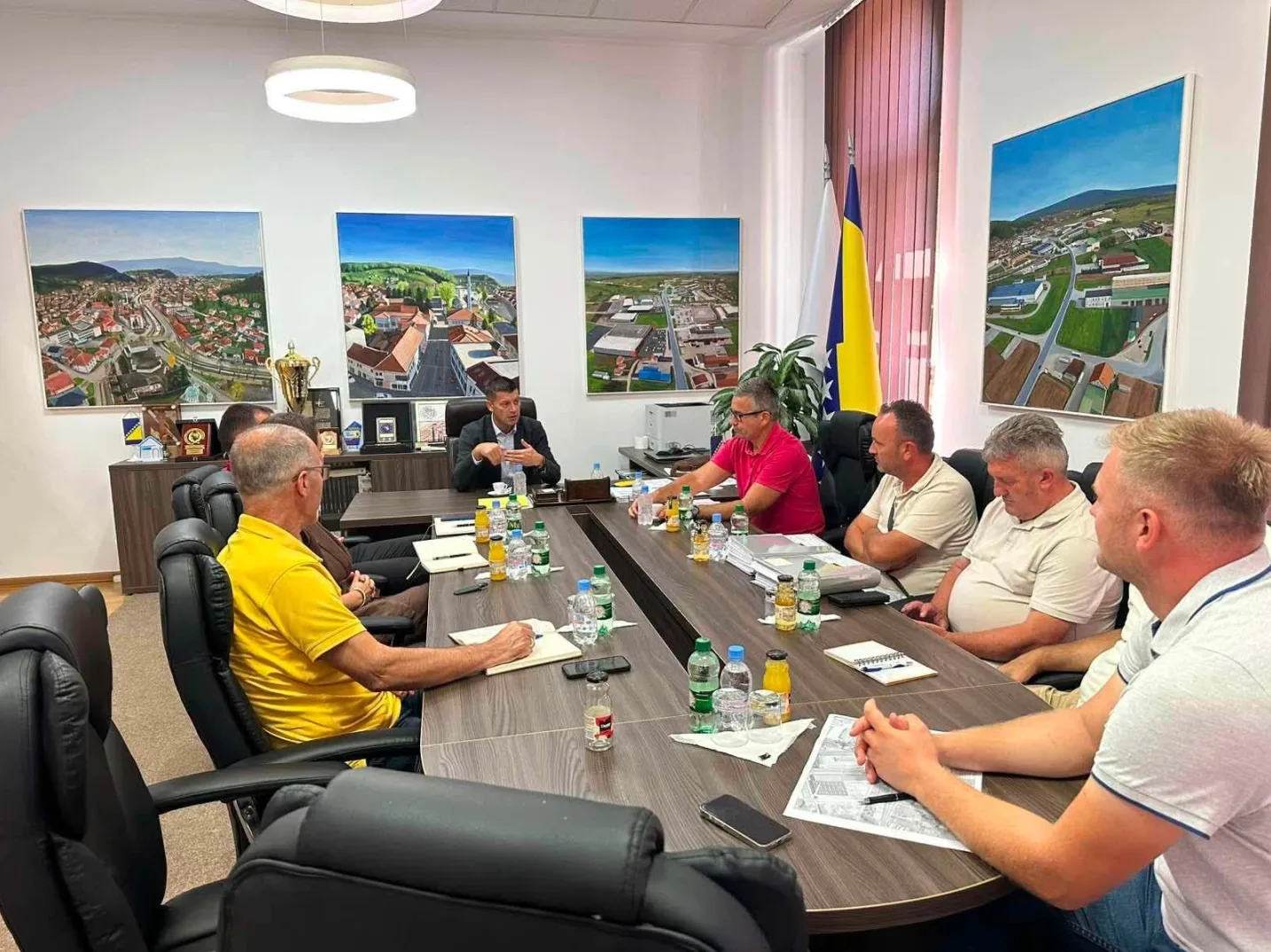 Grupa muškaraca sjedi za velikim stolom na sastanku u modernom kabinetu, s zastavama Bosne i Hercegovine i slikama gradova na zidovima.