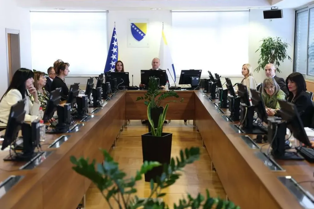 Grupni sastanak u konferencijskoj sali sa ljudima koji sjede za stolom i gledaju u monitore, zastava Bosne i Hercegovine i još jedna zastava u pozadini.
