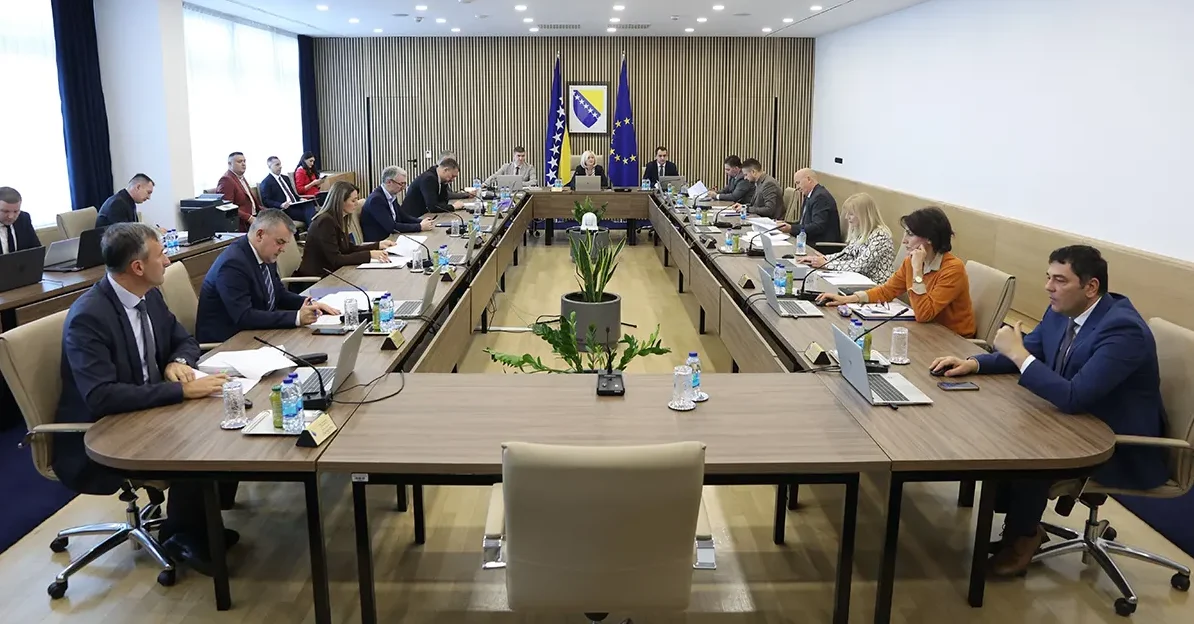 Učesnici sastanka sjede za dugim stolom u modernoj dvorani, ispred njih su laptopi i dokumenti, a na zidu se nalaze zastave Bosne i Hercegovine i Evropske unije.