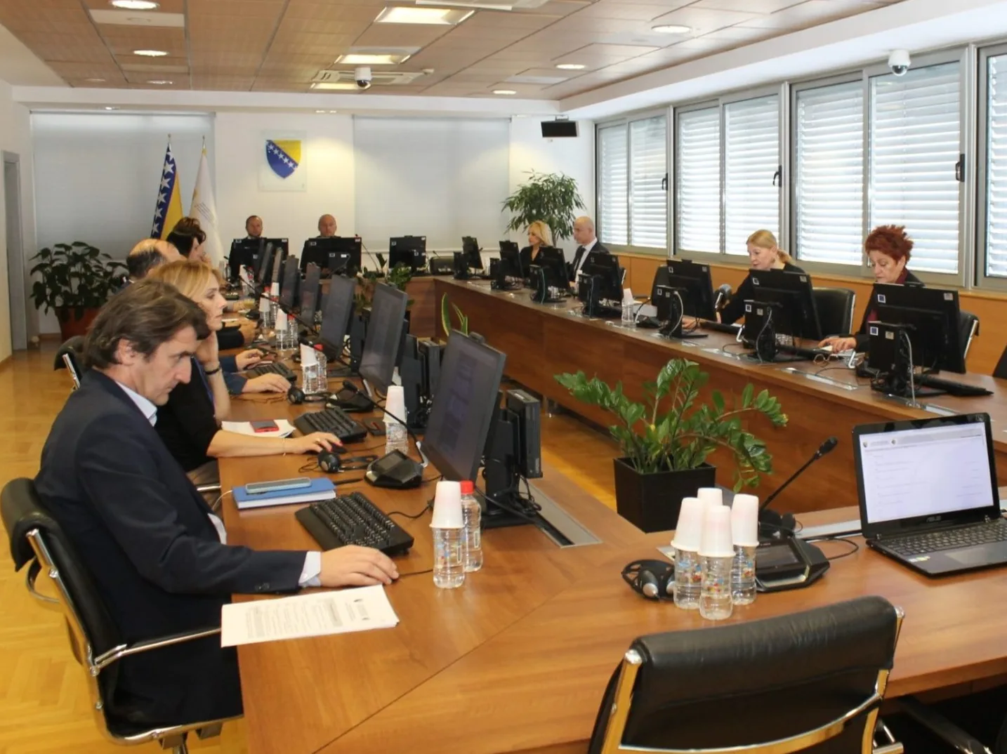 Grupa ljudi sjedi za dugim drvenim stolom u prostoriji za sastanke i radi na računarima.