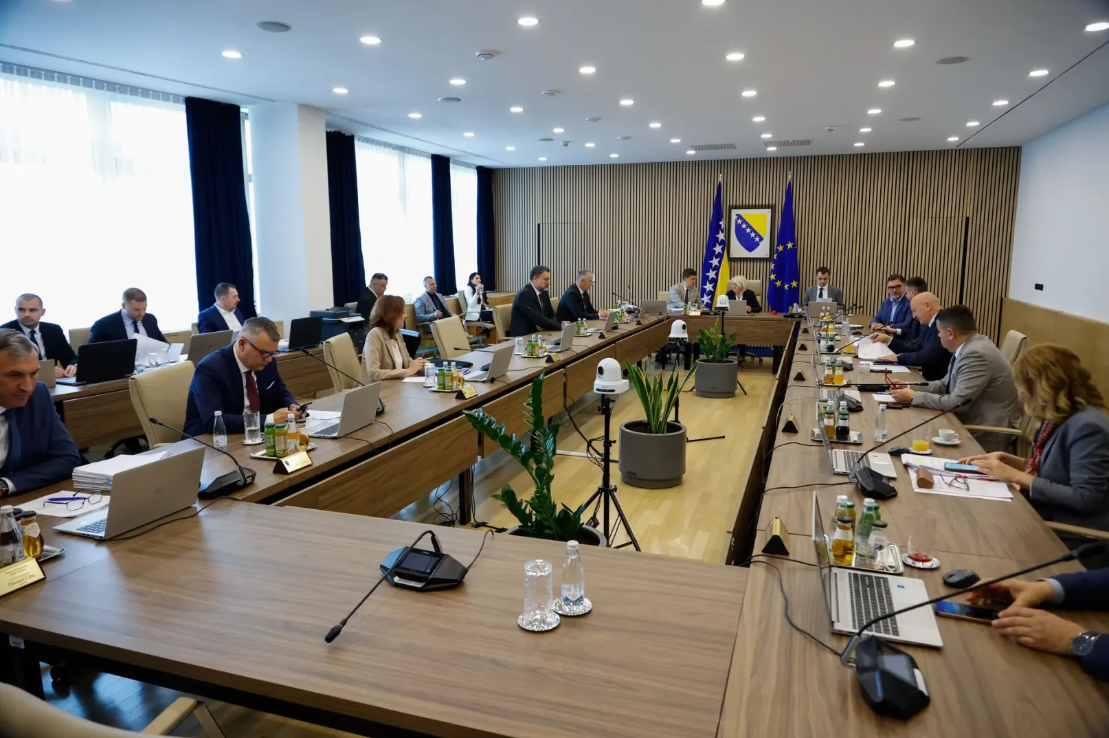 Grupa ljudi sjedi za dugim drvenim stolom u modernoj sali, okružena biljkama i sa zastavama Bosne i Hercegovine i Evropske unije u pozadini.