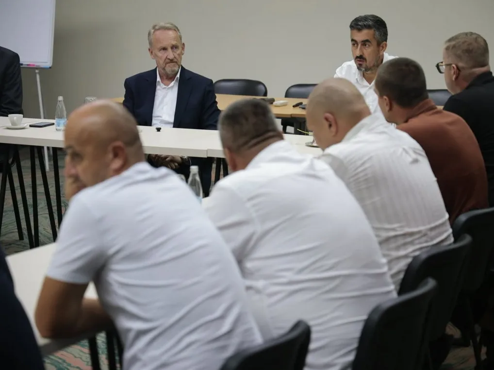 Grupa muškaraca sjedi za stolom na sastanku, fokus na starijem muškarcu u tamnom odijelu i mlađem muškarcu sa naočalama