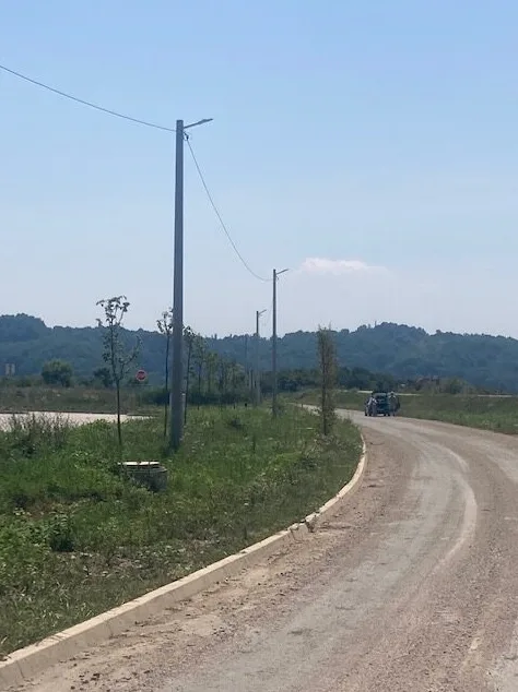 Seoska makadamska cesta krivuda kroz zeleno područje pored stubova javne rasvjete, s drvećem i brdima u pozadini.