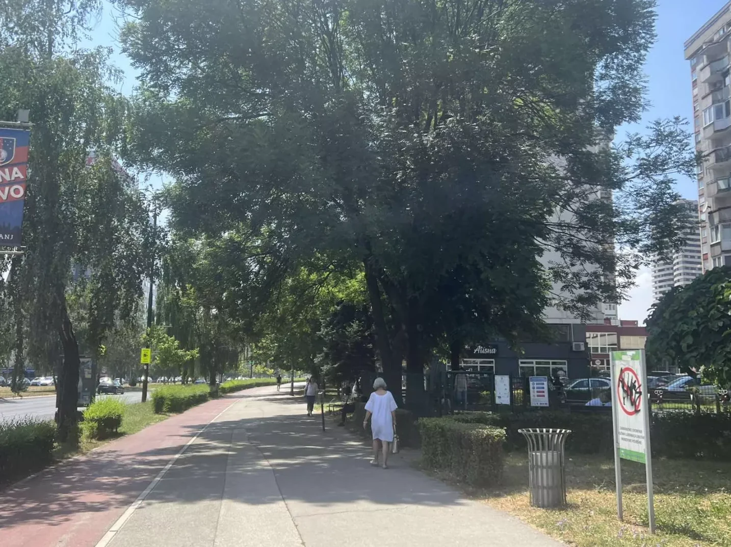 Žena u bijeloj haljini šeta stazom u parku pored drvoreda i zgrada, uz biciklističku stazu.