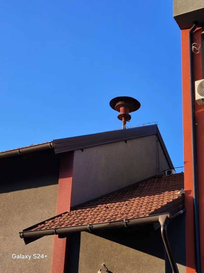 Crvena sirena za uzbunjivanje postavljena na vrhu dimnjaka na krovu zgrade, s jasnim plavim nebom u pozadini.