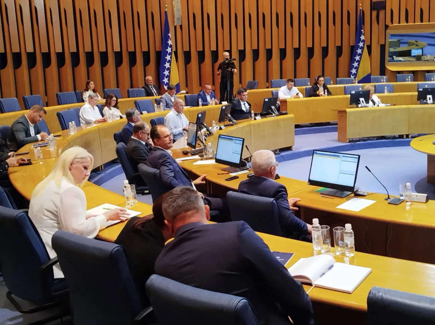 Ljudi sjede za stolovima u sali za sastanke sa zastavom Bosne i Hercegovine