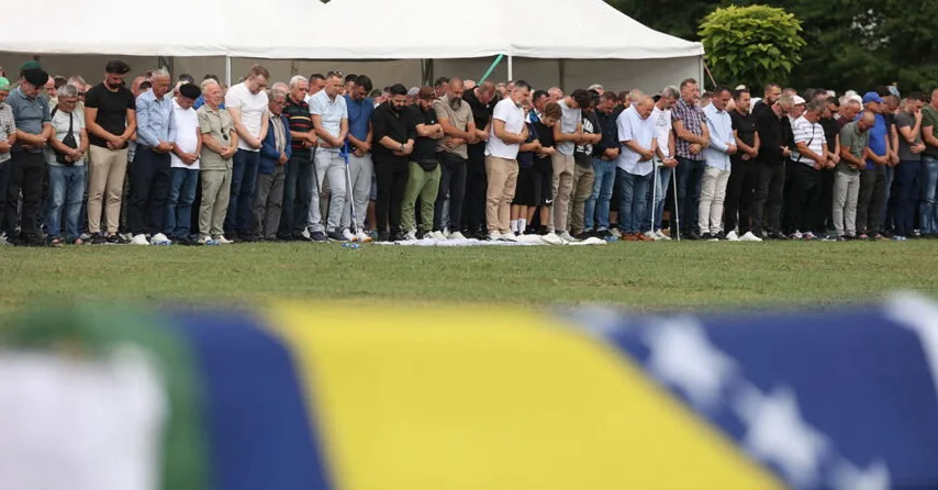 Grupa muškaraca u molitvi na otvorenom, sa zastavom Bosne i Hercegovine vidljivom u prvom planu.