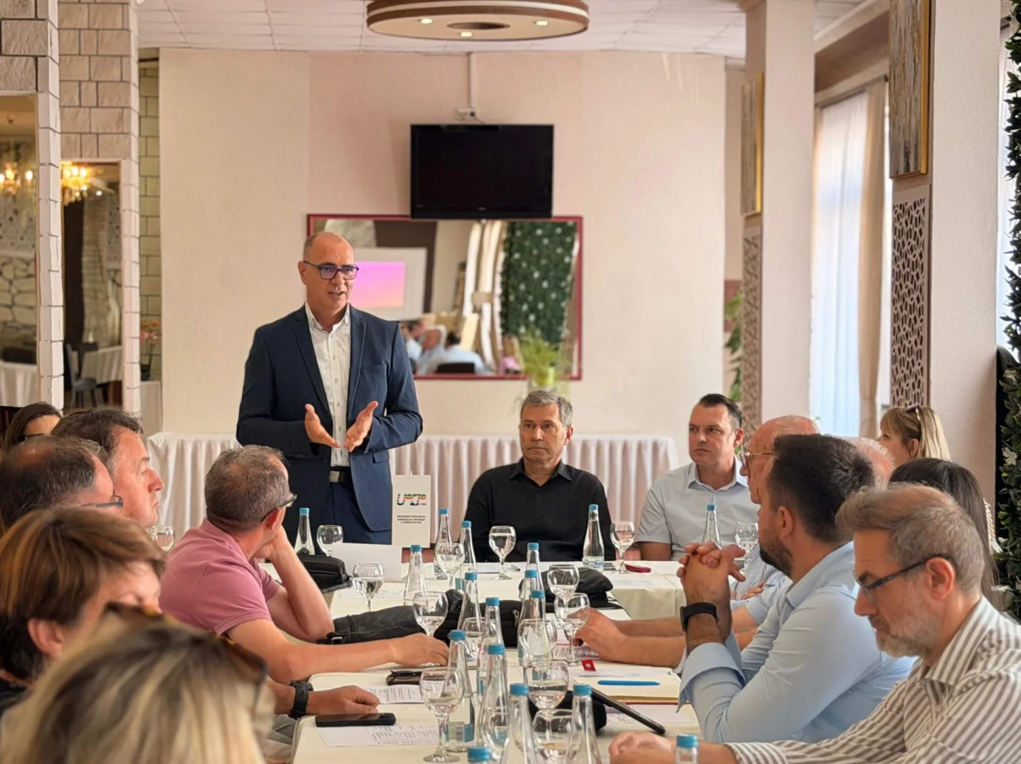 Grupa ljudi sjedi za stolom tokom sastanka, dok im jedan muškarac u tamnom odijelu, naočarima i bijeloj košulji, drži prezentaciju.