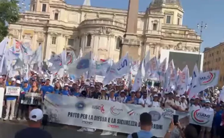 Grupa ljudi na javnom skupu drži brojne zastave i transparent.