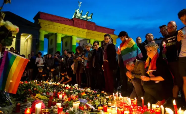 Grupa ljudi pali svijeće i drži dugine zastave ispred Brandenburških vrata u Berlinu, osvijetljenih bojama duge.