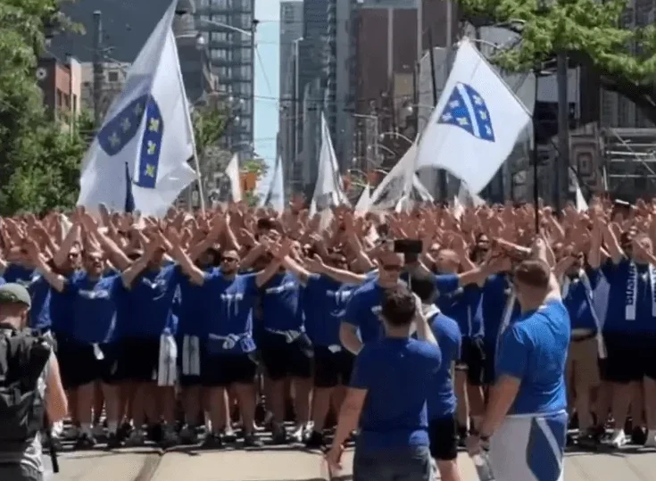 Grupa ljudi u plavim majicama podignutih ruku, drži bijele zastave sa plavim simbolima