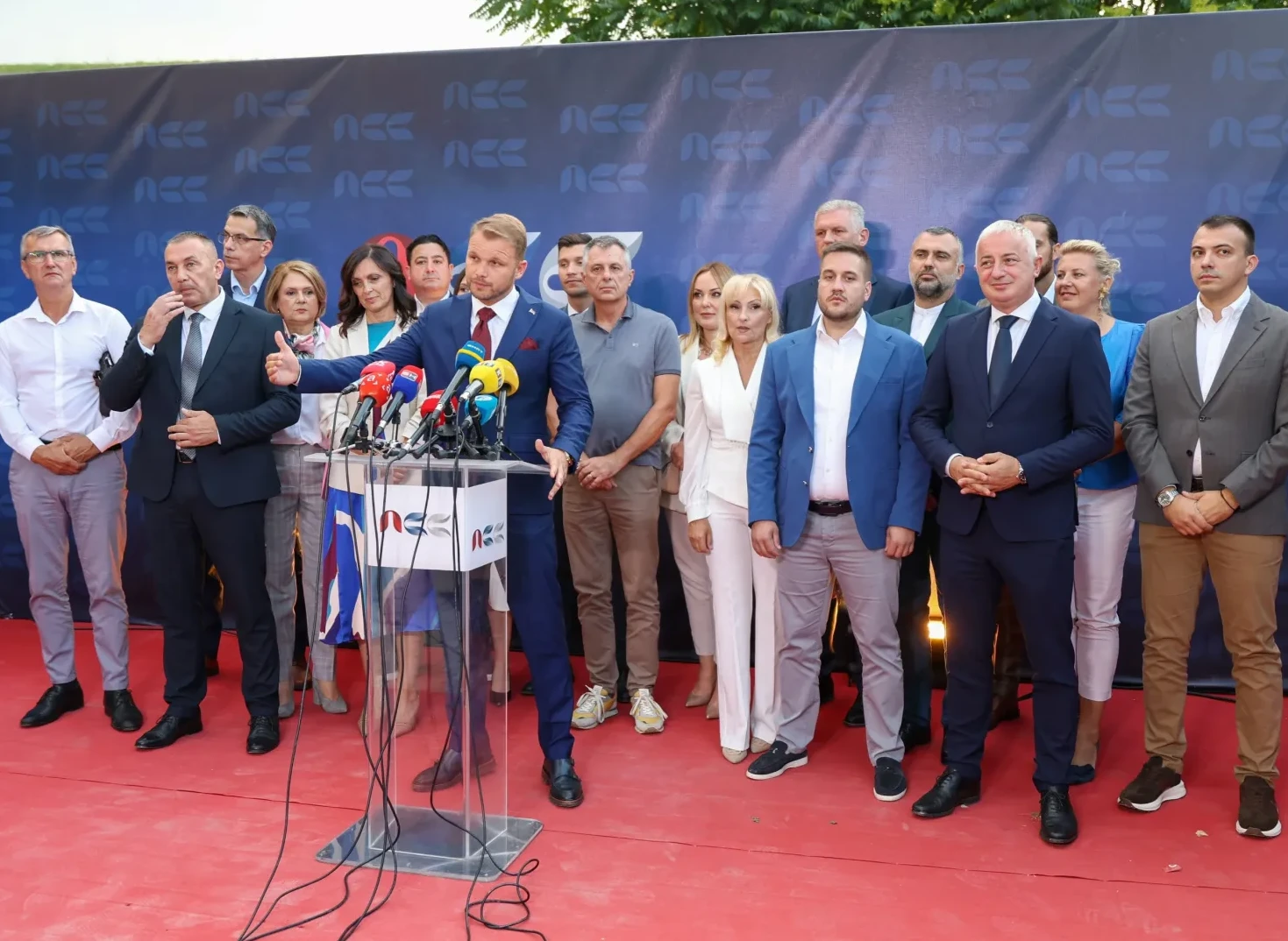 Grupa političara i javnih ličnosti pozira iza govornice sa mikrofonima na crvenom tepihu.