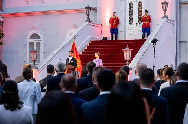 Muškarac u crnom odijelu drži govor za govornicom, pored njega stoji žena u ljubičastoj haljini, a u pozadini se nalaze dva stražara u svečanim uniformama na stepeništu.