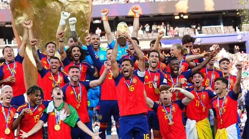 Slika prikazuje slavlje španske nogometne reprezentacije nakon osvajanja trofeja, s igračima odjevenim u crvene dresove, podižući pehar u zrak i noseći zlatne medalje oko vrata.