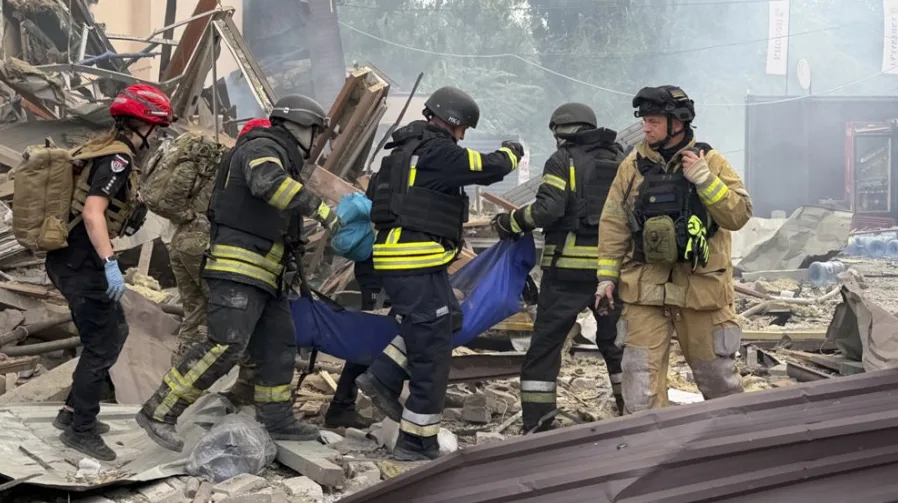 Grupa spasioca u zaštitnim odorama, od kojih neki nose plavo platno, pomno iznose povrijeđenu osobu iz ruševina zgrade nakon eksplozije.