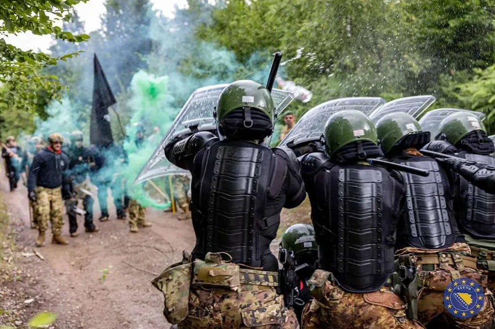 Specijalne jedinice u punoj opremi, noseći prozirne štitove i šljemove, formiraju kordon. U pozadini se vidi dimne bombe zelene boje i crna zastava.