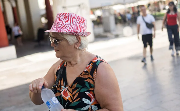 Žena sa kratkom sijedom kosom nosi šešir sa crveno-bijelim zebrastim uzorkom i sunčane naočare. Na sebi ima haljinu sa floralnim motivima. U ruci drži bocu vode. U pozadini se vide nejasne siluete ljudi koji šetaju na sunčan dan.