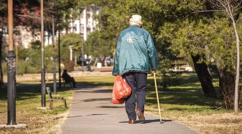 Stariji muškarac u zelenoj jakni i šeširu šeta stazom u parku, držeći štap u desnoj ruci i noseći crvenu plastičnu vrećicu s namirnicama.