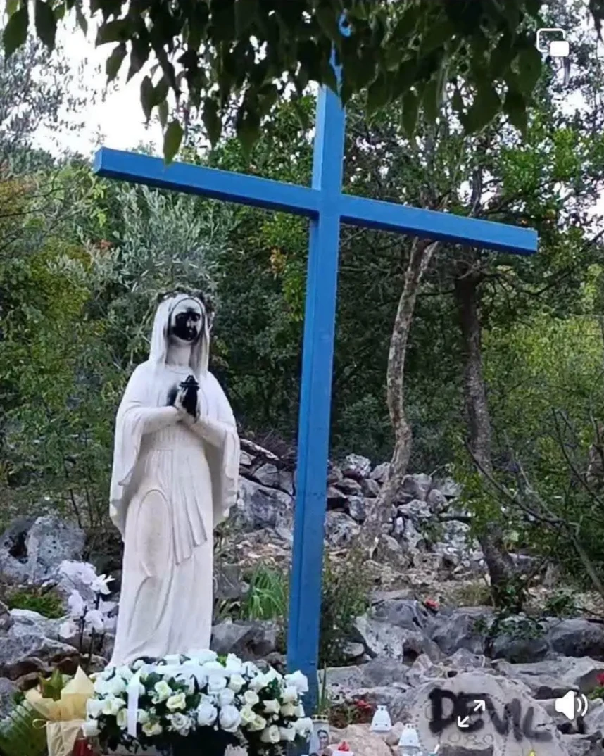 Statua Gospe s rukama sklopljenim u molitvi, pored nje stoji plavi drveni krst. Ispred statue je buket bijelog cvijeća, a u pozadini se vidi kamenjar i drveće. Na jednom od kamena u pozadini ispisano je 'DEVIL'.