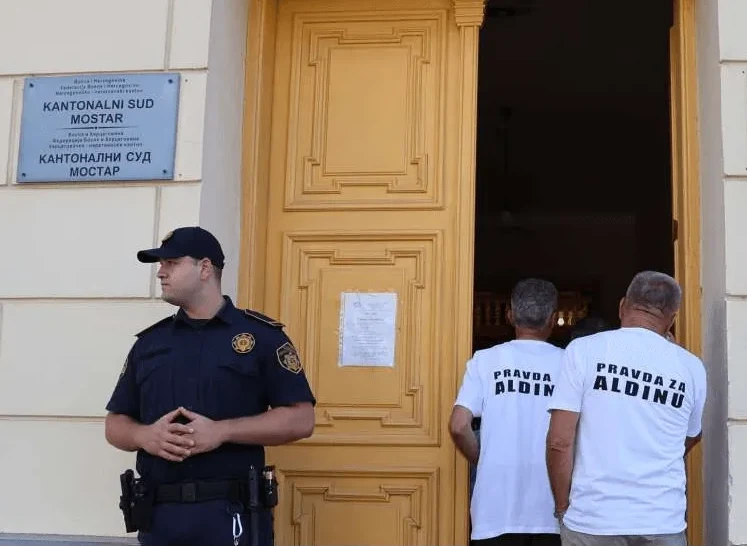 Policajac ispred zgrade Kantonalnog suda Mostar, sa dvojicom muškaraca koji nose majice s natpisom 'Pravda za Aldinu'.