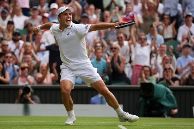 Teniser u bijeloj sportskoj opremi uzvikuje pobjedu i drži reket na travnatom terenu, uz publiku u pozadini na Wimbledonu.