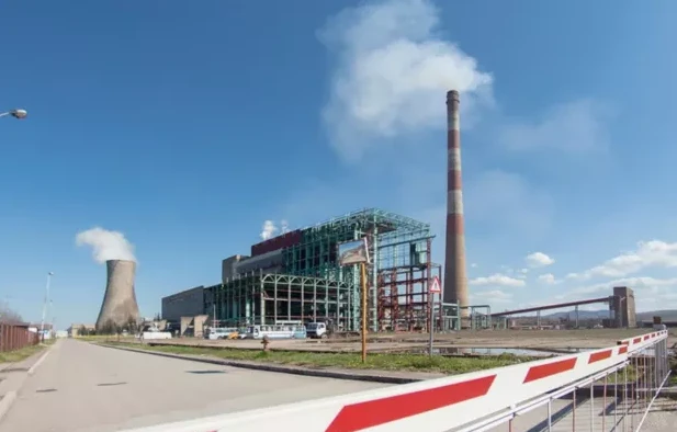 Fotografija prikazuje termoelektranu s rashladnim tornjem iz kojeg izlazi para, te visokim, prugastim dimnjakom iz kojeg također izlazi dim. U pozadini se vidi industrijska postrojenja i ograda s rampom.