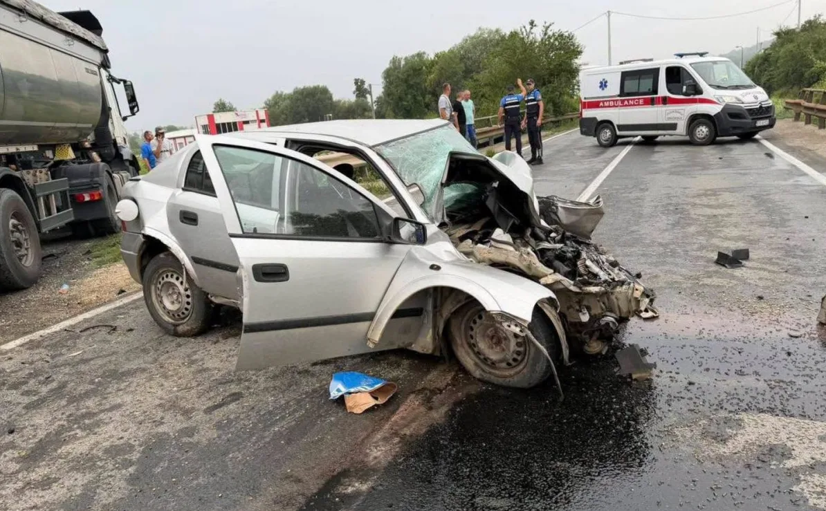 Teško oštećeni srebrni automobil na putu nakon saobraćajne nesreće, pored kamiona i vozila hitne pomoći.