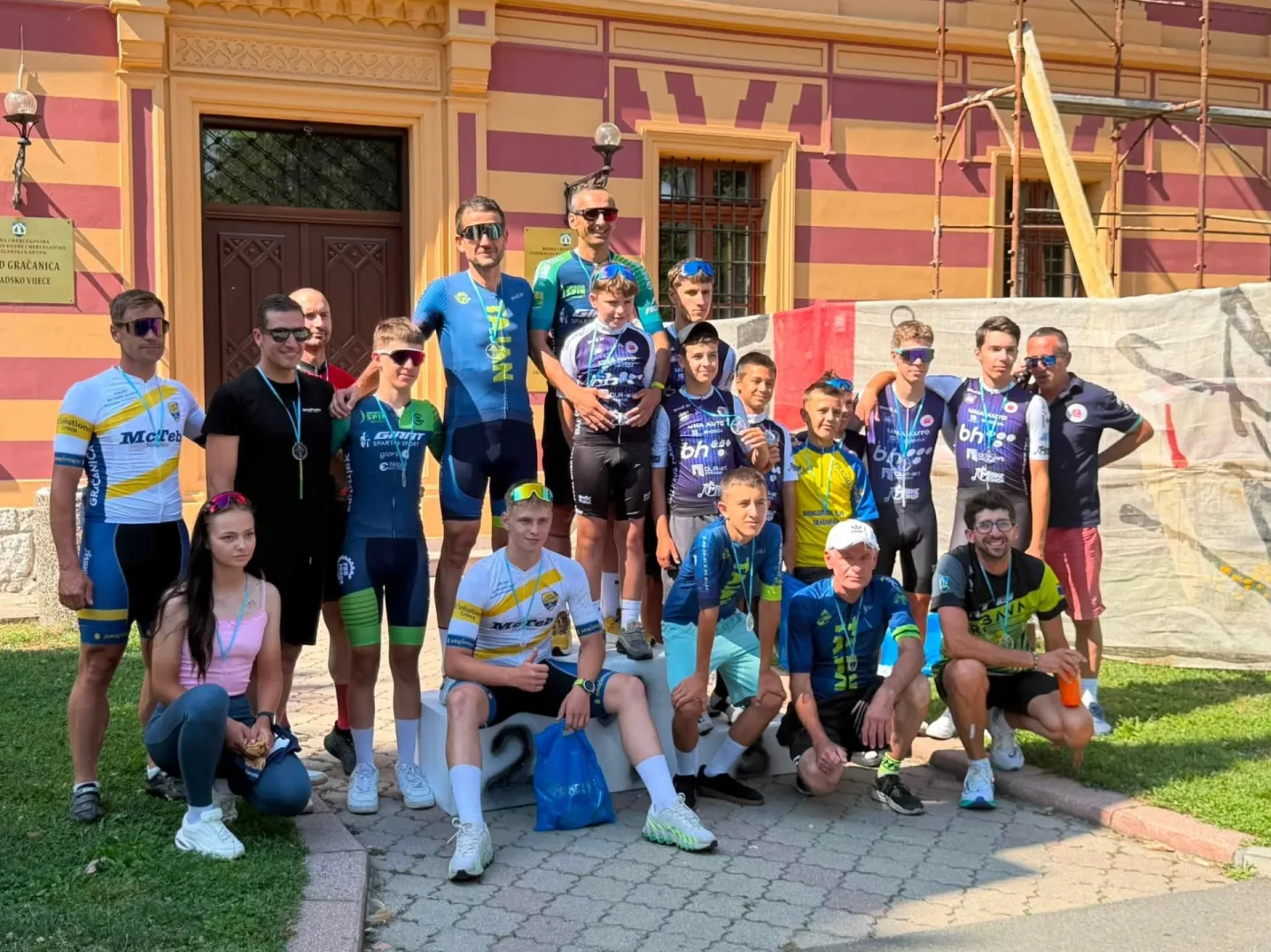 Grupa biciklista, muškarci i jedna žena, poziraju za fotografiju ispred zgrade sa natpisom "Grad Gračanica". Mnogi od njih nose sportske dresove sa logotipima sponzora i medalje oko vrata. Nekolicina mlađih biciklista sjedi na stepenicama, dok ostali stoje. Izgledaju kao pobjednici ili učesnici biciklističkog takmičenja.
