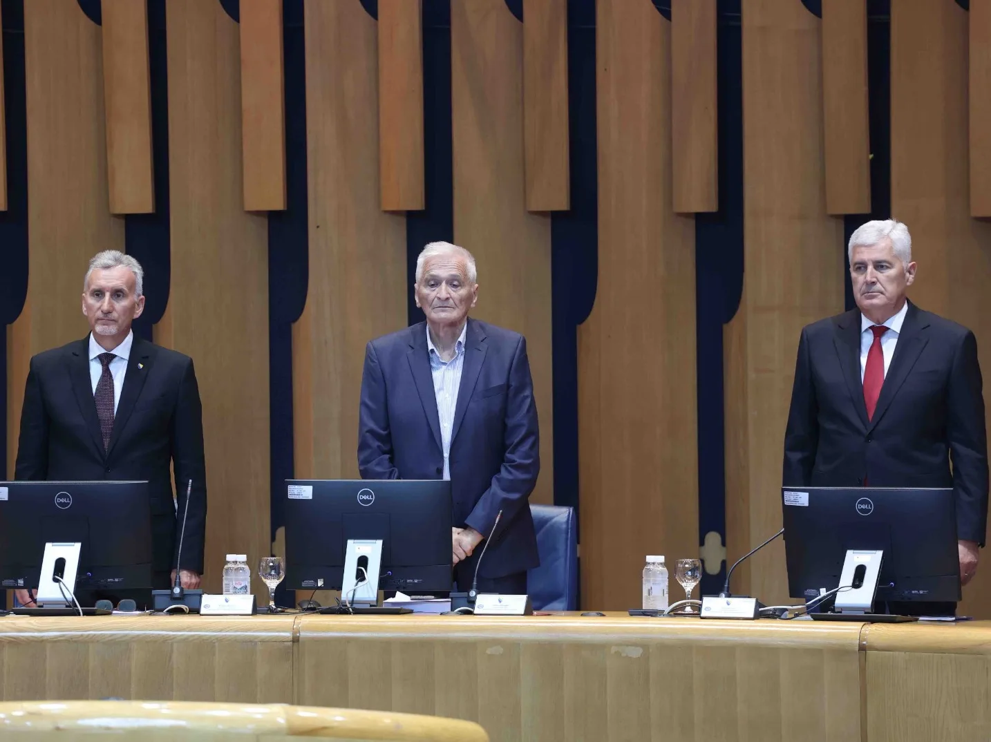 Tri muškarca u odijelima stoje pred računarima u svečanoj sali.