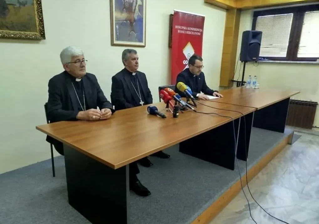 Tri vjerska dostojanstvenika sjede za drvenim stolom na podijumu, ispred njih nalaze se mikrofoni medija i flaše vode. Oni su odjeveni u crne haljine sa bijelim okovratnicima, što ukazuje na njihovu vjersku pripadnost. U pozadini je crveni transparent sa natpisom "Biskupska konferencija Bosne i Hercegovine" te slika na zidu i prozor.
