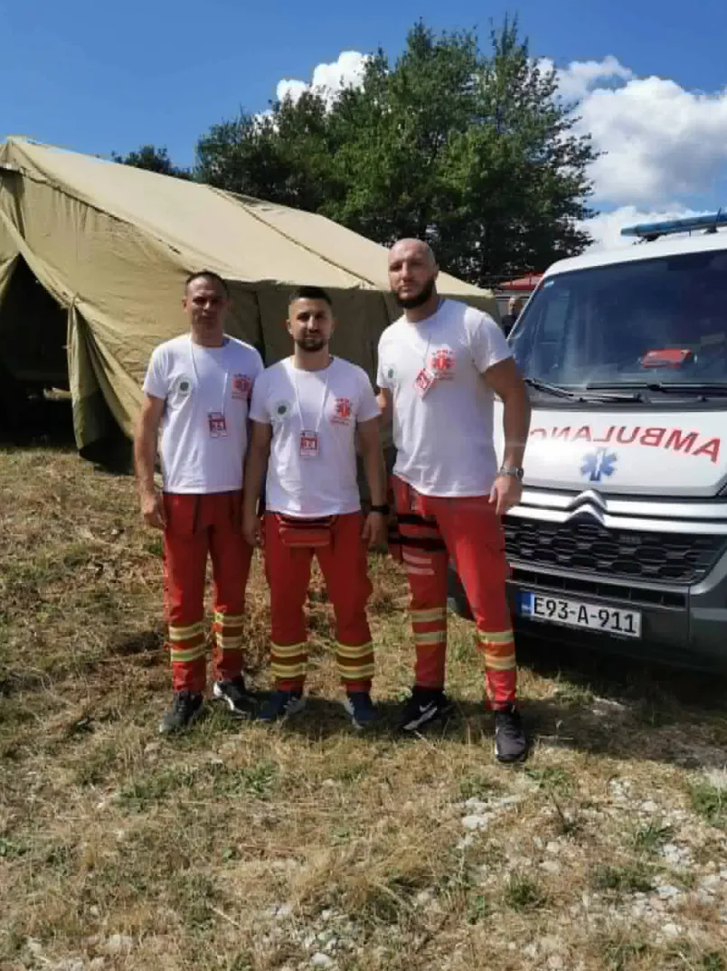 Tri medicinska radnika u crvenim pantalonama i bijelim majicama sa znakom Crvenog krsta, stoje ispred velikog vojnog šatora i vozila hitne pomoći na parkingu.