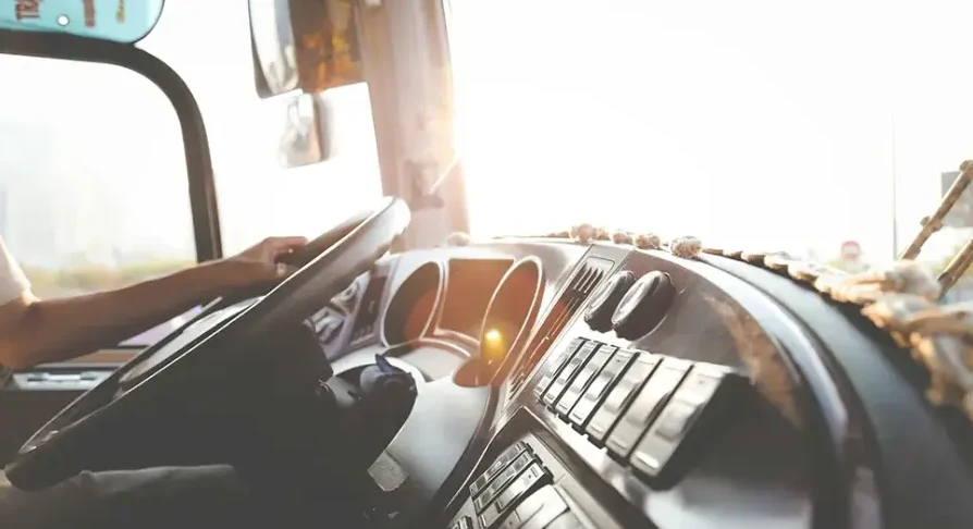 Vozač autobusa drži volan dok sunce zalazi, osvjetljavajući komandnu tablu.