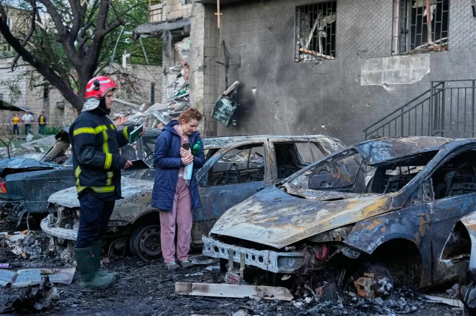 Vatrogasac u zaštitnom odijelu i sa kacigom razgovara sa ženom uplakana lica, okruženi su sa nekoliko potpuno izgorjelih automobila, u pozadini se vidi uništena zgrada sa ostacima vatre i dima.