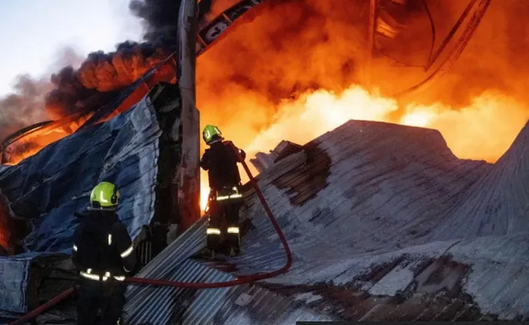 Dva vatrogasca u zaštitnoj opremi i kacigama bore se sa velikim požarom na metalnom skladištu.
